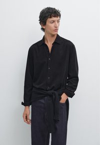 Schwarzes Hemd mit Knöpfen aus weichem Stoff und spitzem Kragen. Hochgerollte Ärmel und ein Pullover um die Taille gebunden. Dunkle Jeans mit Pocket-Detail.