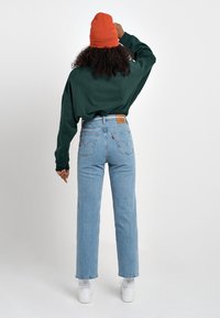 Sudadera verde de gran tamaño, jeans de cintura alta azul claro con un parche de cuero y gorro rojo. Zapatillas blancas completan el atuendo.