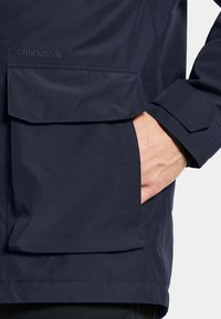 Veste imperméable bleue marine avec une grande poche avant, texture lisse et poignets ajustables. Le nom de la marque "DIDRIKSONS" est embossé sur le tissu.