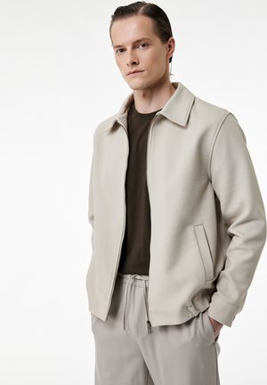 Ung mand iført beige lynlåstrøje over mørk t-shirt og lysegrå bukser, stående med den ene hånd i lommen mod hvid baggrund.