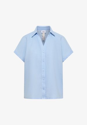 Kvinde med lyst brunt hår iført en lyseblå kortærmet skjorte med knapper og en hvid top, smilende og bærende en beige flettet taske.