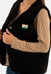 Zwarte fleece vest met ronde kraag, voorzien van beige knopen en een stoffen label met de tekst "ONLY VINTAGE," gedragen over een beige top.