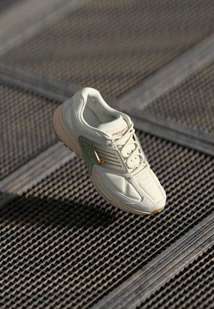 Lys beige og grøn sportsko med beige sål og orange logo hviler på en metalrist.