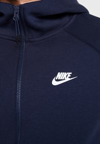 Marinblå zip-up huvtröja i mjukt material, med hög krage och vitt Nike-logotyp på bröstet. Slät textur med atletisk design.