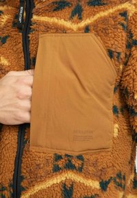 Bruine fleece jas met een gestructureerd patroon, voorzien van een beige borstzak en een rits, met het label "REVOLUTION" nabij de hals.
