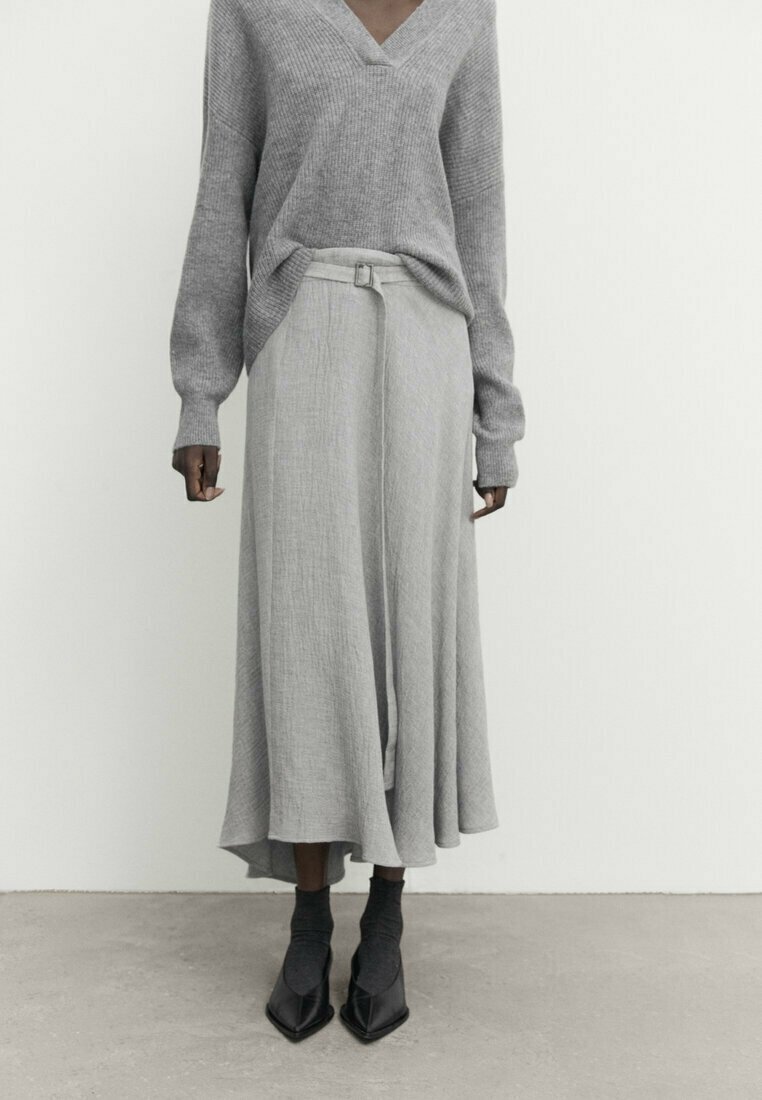 Maglione grigio lavorato a maglia con scollo a V, abbinato a una lunga gonna grigia con cintura e design fluido. Le scarpe nere con punta appuntita completano il look.