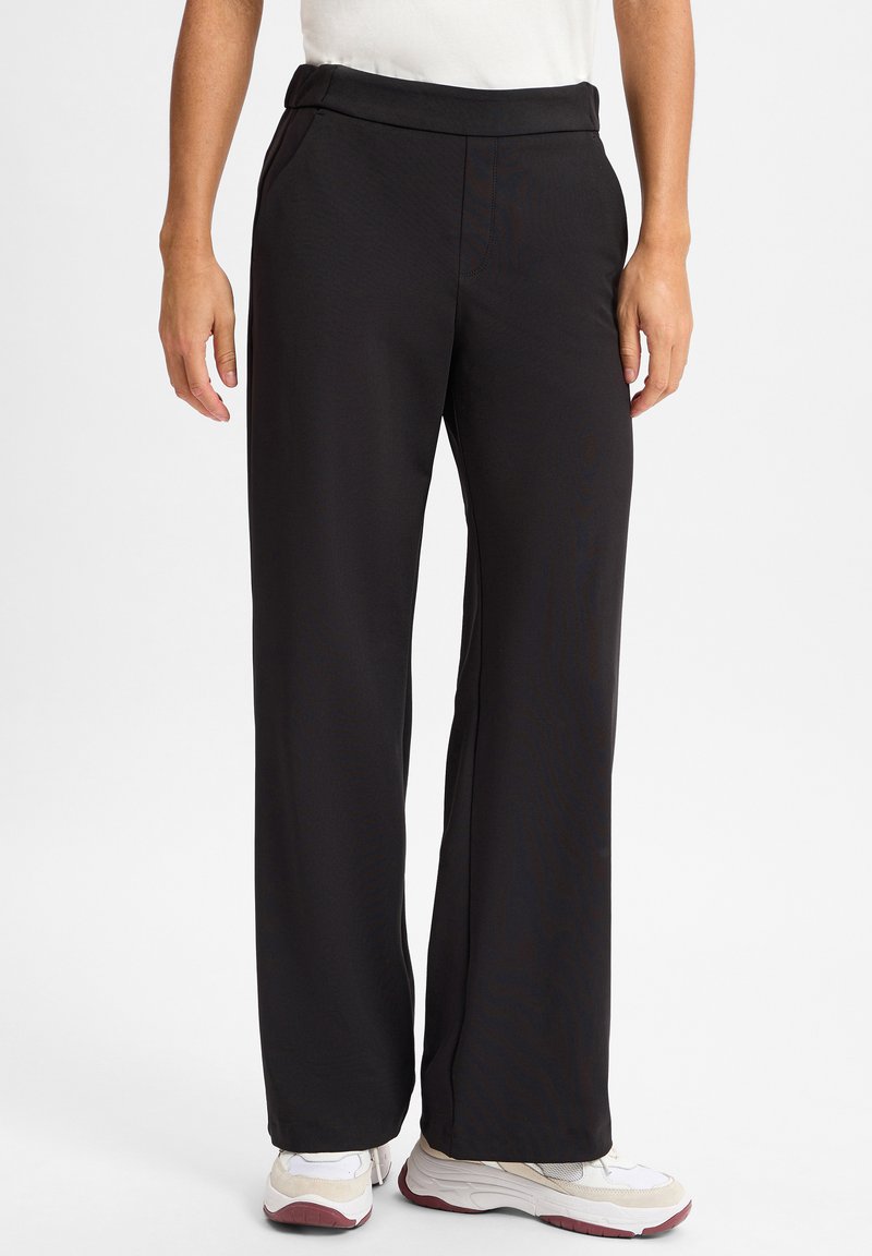 Zwarte, high-waisted broek met een gladde textuur, recht model, zijzakken en een subtiele glans. Gestyled met witte sneakers.