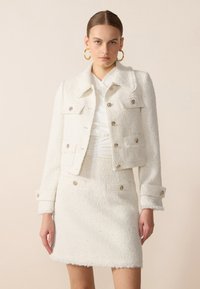 Giacca bianca in tweed con colletto e tasche frontali, abbinata a una gonna coordinata con orlo sfilacciato e bottoni metallici decorativi.