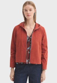 Veste rouge à capuche en matière légère, dotée d'une fermeture éclair à l'avant, de deux poches latérales et d'une coupe courte. Portée sur un haut à motifs floraux.