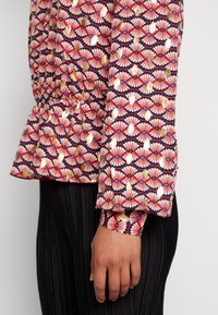 Bluse mit floralem Muster in Rot-, Blau- und Goldtönen. Mit einer gerafften Taille und eng anliegenden Bündchen. Das strukturierte Material erhöht die visuelle Detailtiefe.