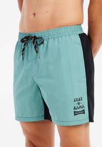 Turquoise en zwarte zwembroek met een elastische tailleband, trekkoord, aan de voorkant een zak en het bedrukte "SURF-O-RAMA" logo. Glad stofmateriaal.
