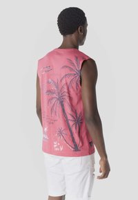 Uomo con canotta rosa senza maniche con stampa di palme nere e pantaloncini bianchi, in piedi di schiena contro uno sfondo uniforme.
