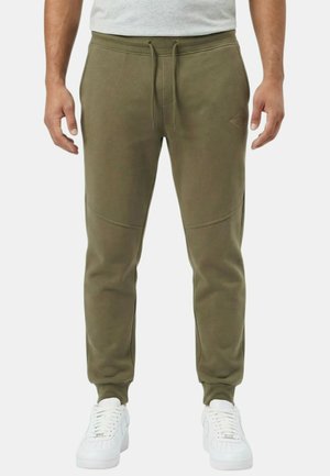 Homme portant un pantalon de jogging vert olive avec une taille à cordon de serrage et des baskets blanches, debout devant un fond clair uni.