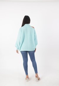 Pullover oversize azzurro chiaro in maglia con collo alto, dettagli spalle scoperte e texture a coste, indossato con jeans blu aderenti e tacchi trasparenti.