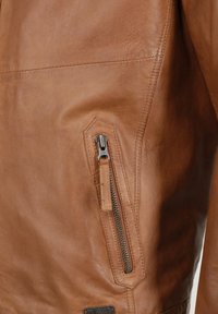 Bruine leren jas met een ritszak en metalen hardware, gladde textuur en een op maat gemaakte pasvorm, benadrukkend vakmanschap en detail.