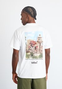 Witte T-shirt met korte mouwen met een kleurrafische afbeelding van een stadsgezicht met een cartoonachtig personage, vergezeld van tekst aan de onderkant.