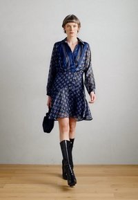 Tamsiai mėlynos spalvos raštuota suknelė su ilgomis rankovėmis, įliemenuota talija ir plačiu sijonu; derinama su juodomis aukštomis batais iki kelių ir maža rankine.