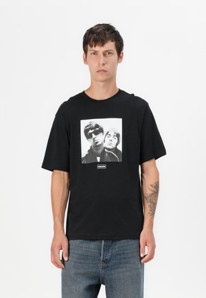 Black cotton t-shirt featuring a central graphic print of two figures in monochrome, with an "oasis" label at the bottom. Short sleeves.