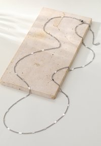 Srebrny naszyjnik łańcuszkowy na beżowej kamiennej powierzchni. Łańcuszek składa się z płaskich, prostokątnych ogniw o gładkiej fakturze oraz zapięcia.