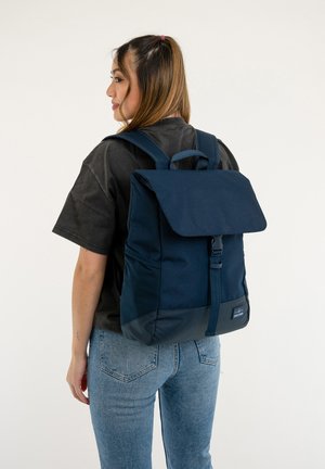 Navy fabric backpack with a flap closure, adjustable straps, and a top handle. Features a textured bottom panel and a logo tag.