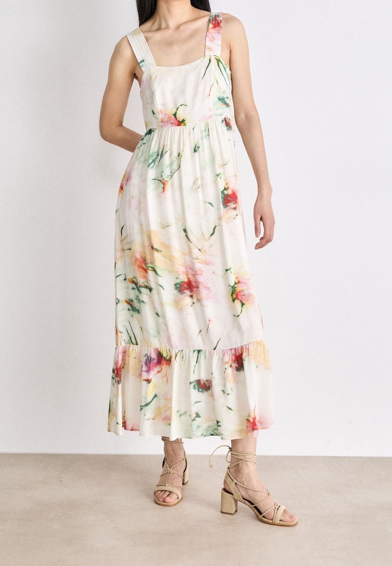 Femme portant une robe midi blanche sans manches avec des imprimés floraux colorés et des sandales beiges à talons blocs lacées, debout devant un fond uni.