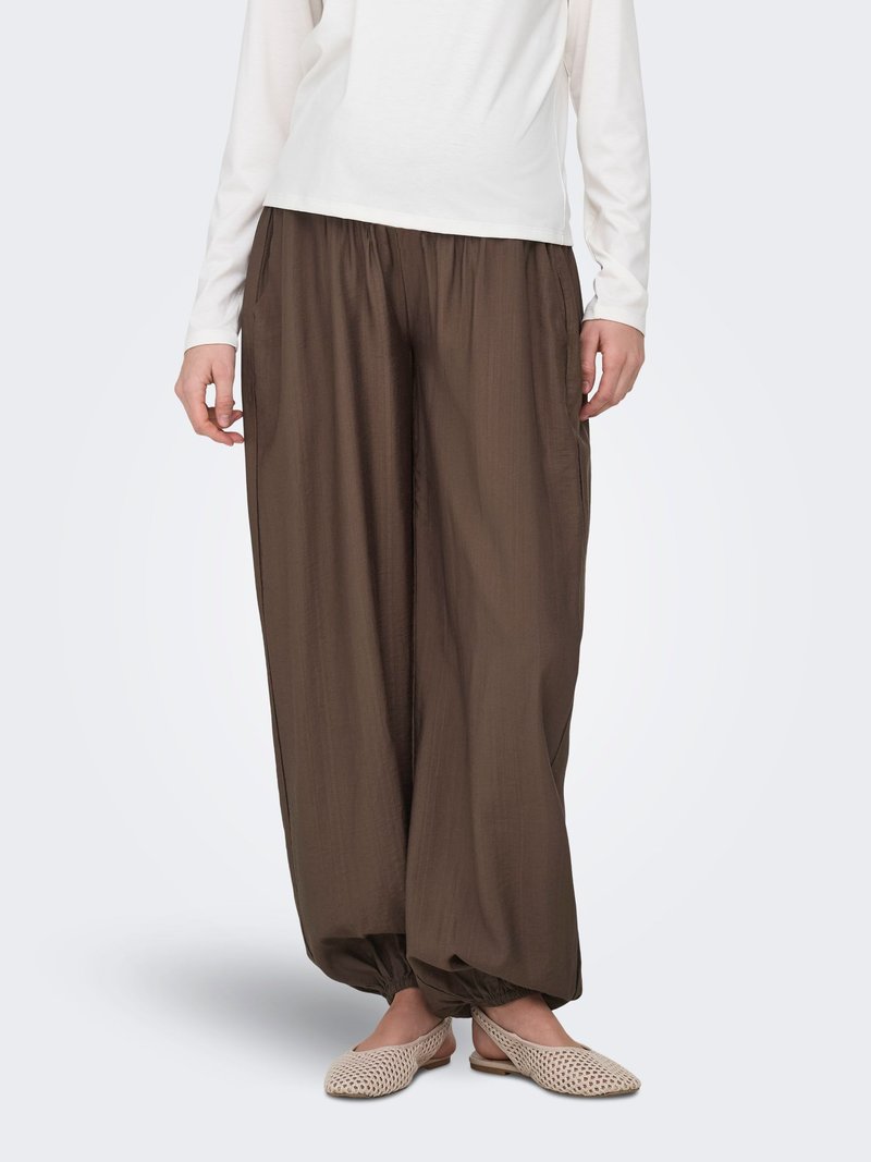 Personne portant un pantalon ample marron avec des poignets élastiques, un haut blanc à manches longues et des chaussures plates tissées beiges, debout devant un fond uni.