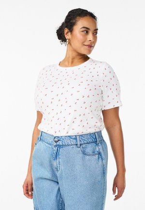 Žena v bielom tričku s krátkym rukávom s malou potlačou červených čerešní a svetlomodrých džínsov s vysokým pásom, ktorá sa pozerá do strany s vlasmi stiahnutými do drdola.