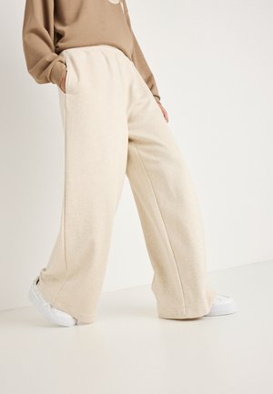 Beżowe spodnie o szerokich nogawkach, wykonane z miękkiego materiału, z bocznymi kieszeniami i elastycznym paskiem w talii, połączone z białymi trampkami na jednolitym tle.