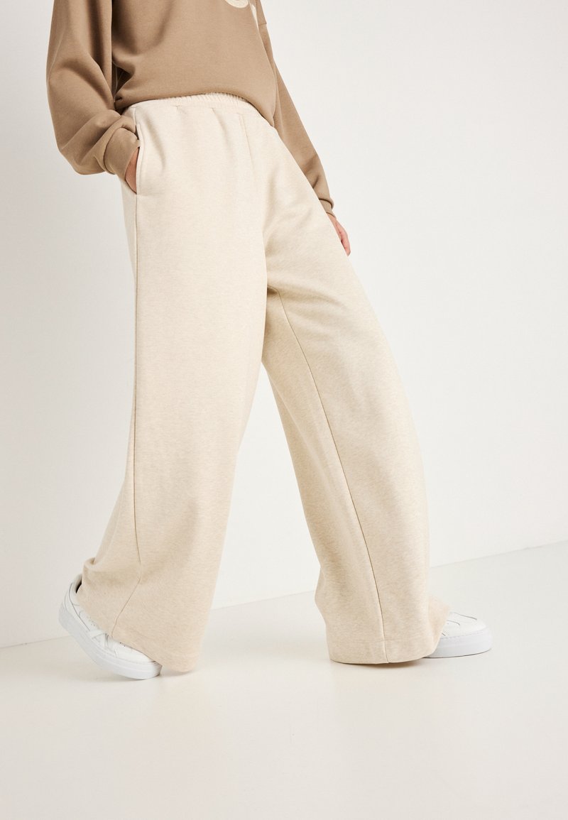 Μπεζ παντελόνια με φαρδιά πόδια φτιαγμένα από μαλακό ύφασμα, με πλαϊνές τσέπες και ελαστική μέση, συνδυασμένα με λευκά αθλητικά παπούτσια σε λείο φόντο.