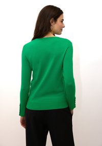 Pull en maille verte avec encolure ronde, manches longues, poignets côtelés et petits boutons décoratifs sur les ourlets des manches. Porté avec un pantalon foncé.