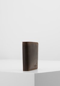 Braune Lederbörse mit einem minimalistischen Design, glatter Oberfläche, sichtbaren Nähten und geprägtem Logo, aufrecht auf einer weißen Oberfläche stehend.
