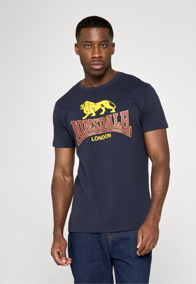 Άνδρας που φοράει μπλε-ναυτικό μπλουζάκι Lonsdale London με κίτρινο λογότυπο λιονταριού, συνδυασμένο με μπλε τζιν, στέκεται μπροστά σε απλό φωτεινό φόντο.