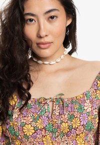 Vestido de estampado floral con escote cuadrado, tela fruncida y detalle de lazo. Presenta colores vibrantes: púrpura, amarillo y verde. Accesorio de collar de conchas.