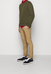 Olivgrön sweatshirt med rund hals, lager på lager över en röd skjorta, tillsammans med beige slim fit-byxor och svarta sneakers med vita sulor.