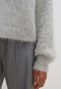 Hellgrauer, flauschiger Pullover mit lockerem Schnitt und breiten Manschetten, kombiniert mit grauen Hosen. Der Pullover hat eine weiche, strukturierte Oberfläche.