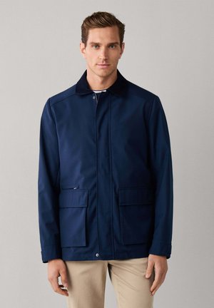 Homme face à l’avant portant une veste bleu marine avec un col noir et un pantalon beige sur un fond gris uni.