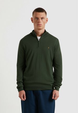Pull-over en maille vert foncé avec col à demi-fermeture éclair et poignets côtelés. Présente un petit logo doré sur la poitrine. Le modèle porte un pantalon bleu marine.