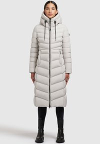 Ljusgrå, lång pufferjacka med huva, med horisontell sömnad, framdragkedja och sidofickor. Bärs med svarta byxor och stövlar.