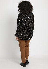 Black long-sleeve blouse with a repeating pattern, paired with brown fitted pants and black footwear. Smooth fabric with a loose fit.