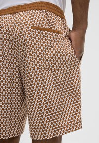 Pantaloni corti a righe marroni e bianchi con vita elastica e tasca posteriore, indossati da una persona con una mano nella tasca laterale.