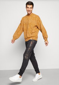 Sennepsfarvet sweatshirt med et struktureret mønster, løs pasform og ribbede manchetter, parret med sorte joggers med blanke sidepaneler og hvide sneakers.