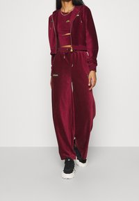 Burgundowy dres z weluru, składający się z krótkiej bluzy z suwakiem i szerokich spodni z elastycznym pasem. Widoczne są złote akcenty oraz detale z logo.