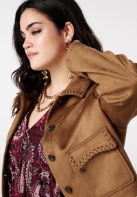 Veste en daim marron avec fermeture à boutons, détail de poche cousue et texture douce, portée sur un haut bordeaux à motifs.