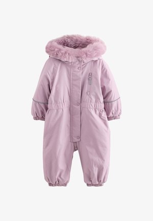 Niño pequeño sonriente con un mono de invierno de color púrpura claro con capucha con borde de piel y botas con estampado floral, de pie sobre una plataforma blanca curva.