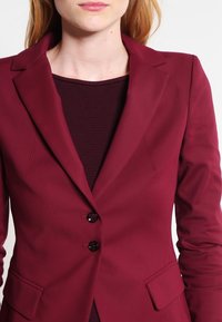 Blazer burdeos con un diseño entallado, dos botones frontales y bolsillos laterales. Tela suave con un ligero brillo y solapas ajustadas.