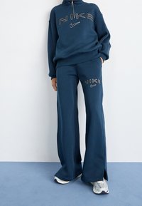 En person som bär en mörkblå Nike-hoodie och vida sweatpants med logotyper, står på ett ljust blått golv mot en vit vägg.