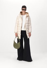Lichtbeige pufferjack met een capuchon, gecombineerd met een witte top en zwarte flared broek. Houdt een gebogen groene handtas vast en draagt donkere zonnebril.