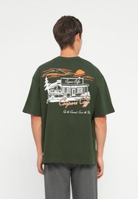 Camiseta de gran tamaño en verde oscuro con una ilustración gráfica en blanco y naranja de un café, con un sol, árboles y un coche vintage.