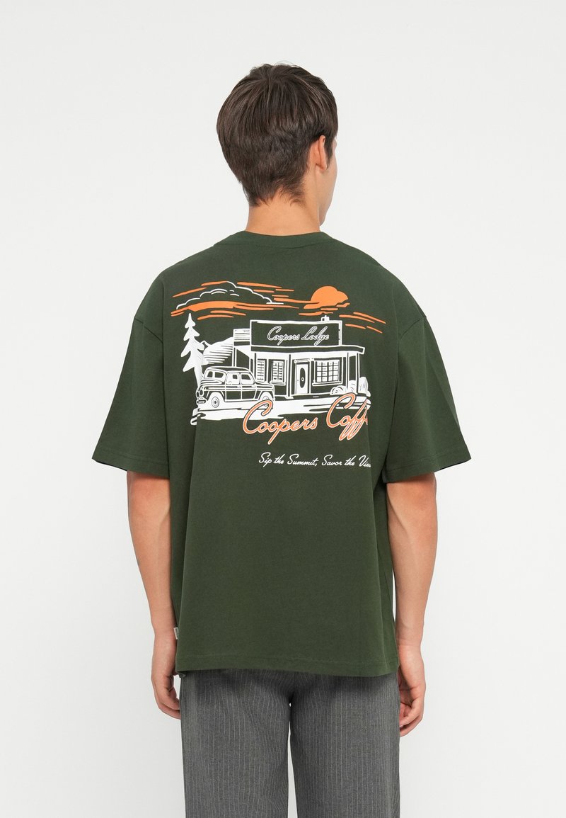 Camiseta de gran tamaño en verde oscuro con una ilustración gráfica en blanco y naranja de un café, con un sol, árboles y un coche vintage.