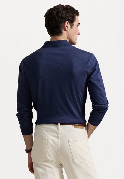 Navyblå långärmad polo med krage, slät textur och figursydda ärmar. Bärs med ljusa byxor och ett brunt bälte.